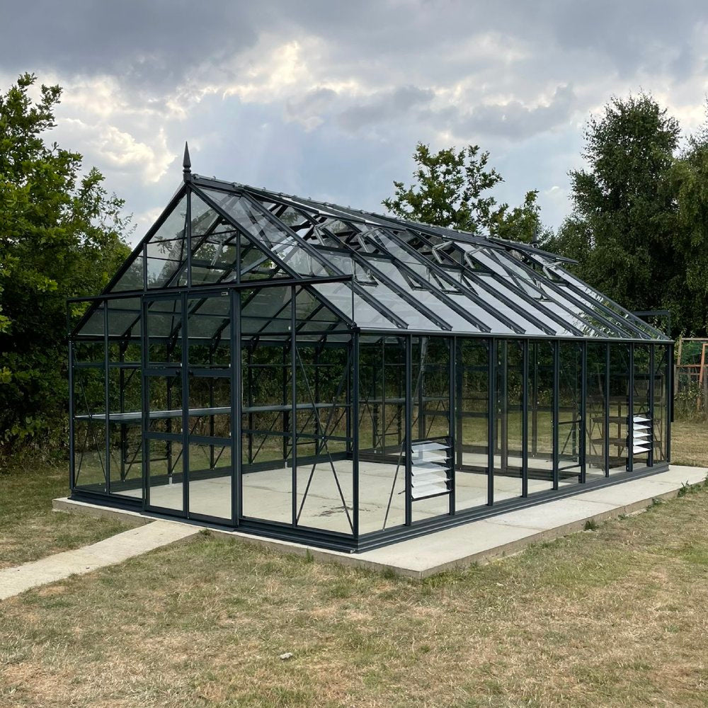 Glass Rhino Greenhouse with a pointed roof stands on a concrete slab, surrounded by grassy field and trees under a cloudy sky.