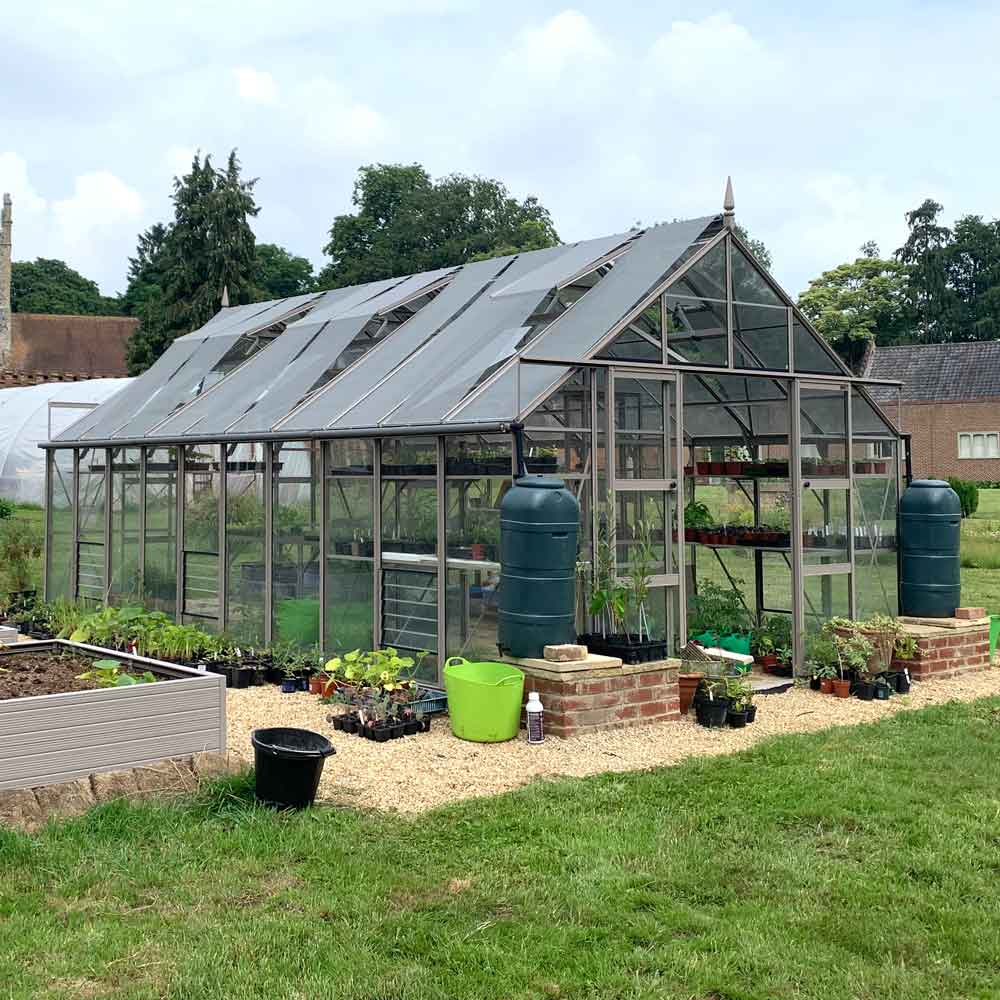 A glass and metal Rhino Greenhouse houses plants in a garden, surrounded by potted plants, green watering can, black bucket, and two blue water tanks, with grassy and landscaped surroundings.