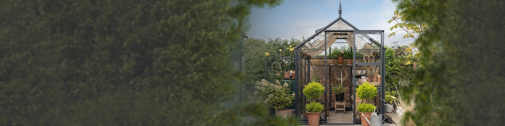 A glass Rhino Greenhouse houses potted plants on shelves, surrounded by garden foliage. Clear sky and trees form the background, enhancing the serene, natural setting.