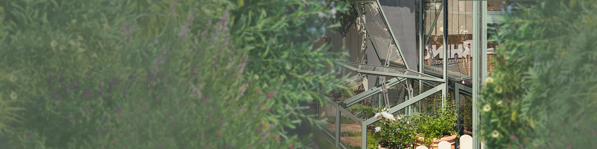 A Rhino Greenhouse with open panels, surrounded by lush plants. Inside, potted flowers are visible. The partially obscured text reads, RHINO Rhino Greenhouses.