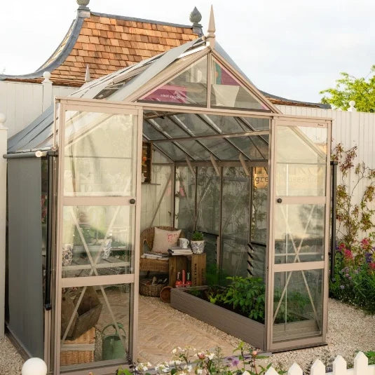 Rhino Greenhouse with open doors displaying potted plants and gardening tools inside, set against a white fence with a wooden-roofed structure nearby, surrounded by a landscaped garden.